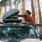 A man loads camping gear into a roof-top cargo carrier attached to his vehicle. 